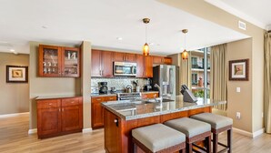 A modern kitchen with wooden cabinets, a central island with bar stools, stainless steel appliances, and pendant lighting. - Maui Resort Rentals: Honua Kai Konea 239 - Upgraded Ocean View 2BR, Deluxe Corner Suite (Lahaina)