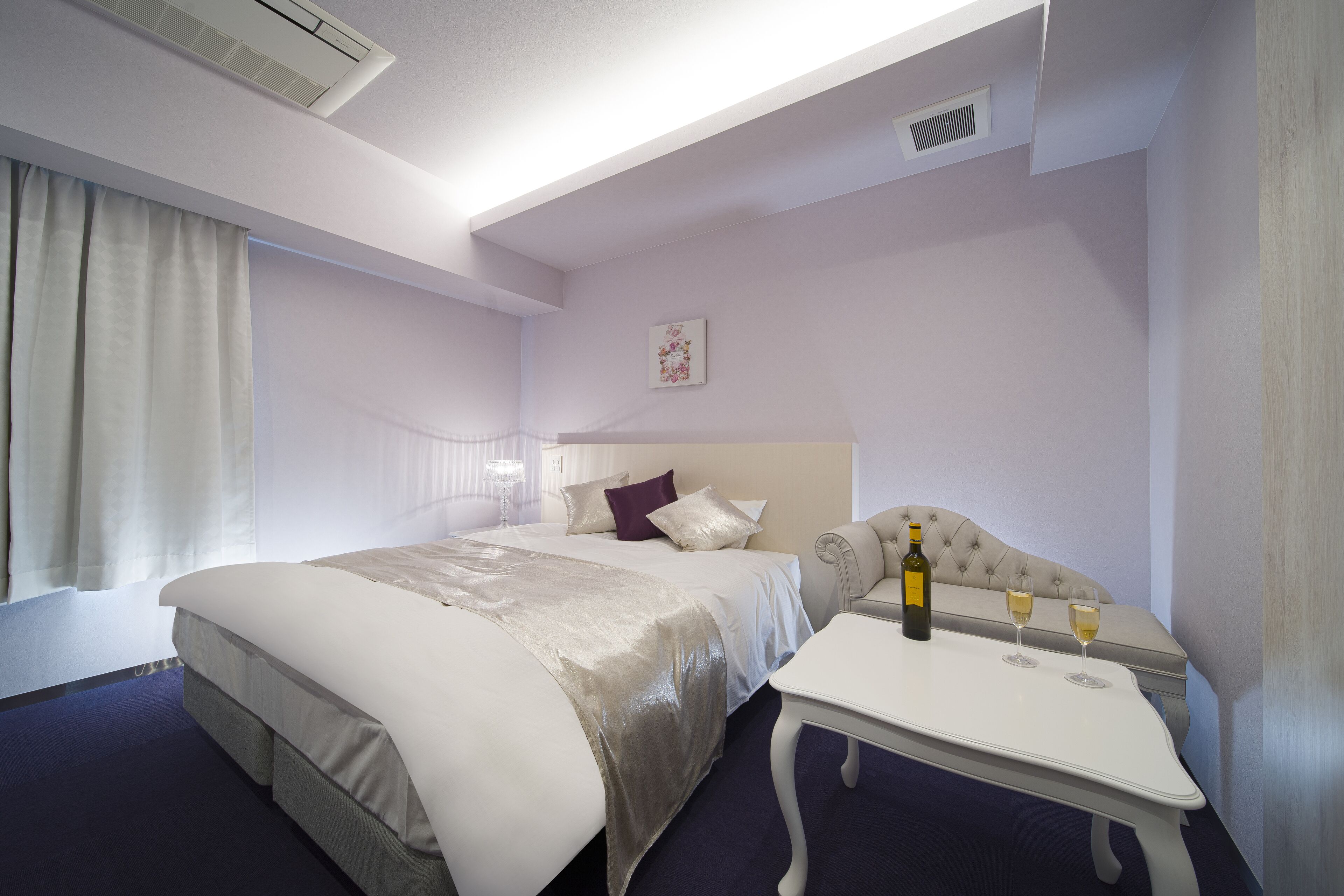 A hotel room with a bed, a bedside table with a bottle and glasses, a window with curtains, and a ceiling vent.