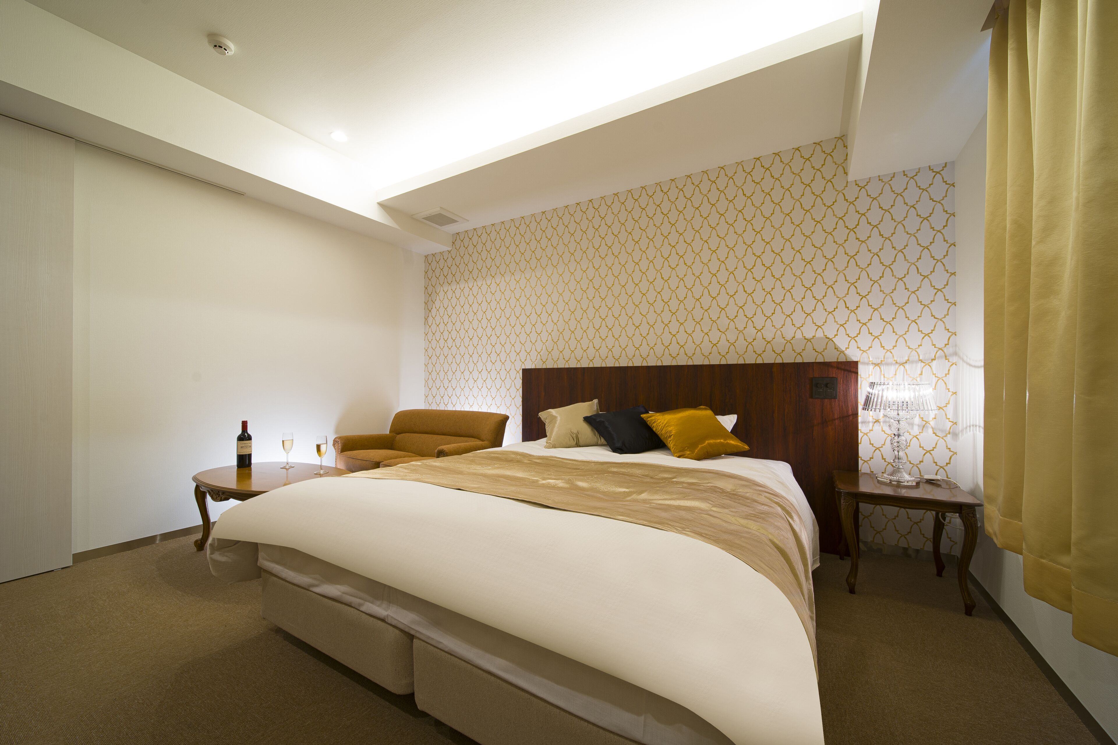 Een hotelkamer met een groot bed, een stoel, een klein tafeltje met een fles en glazen, en een nachtkastje met een lamp.