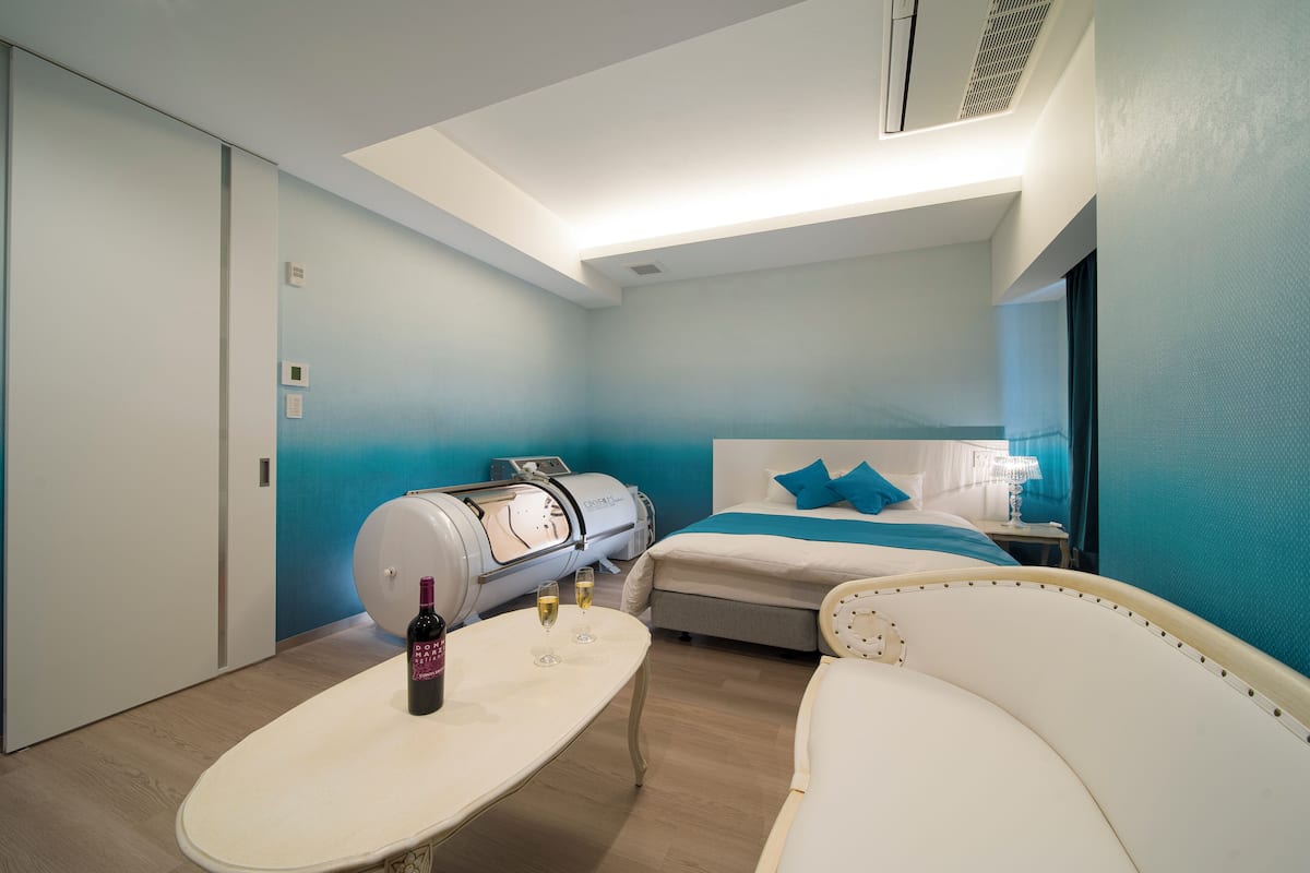 A modern hotel room with a bed, a massage chair, a small table with a bottle and glasses, and a ceiling light.
