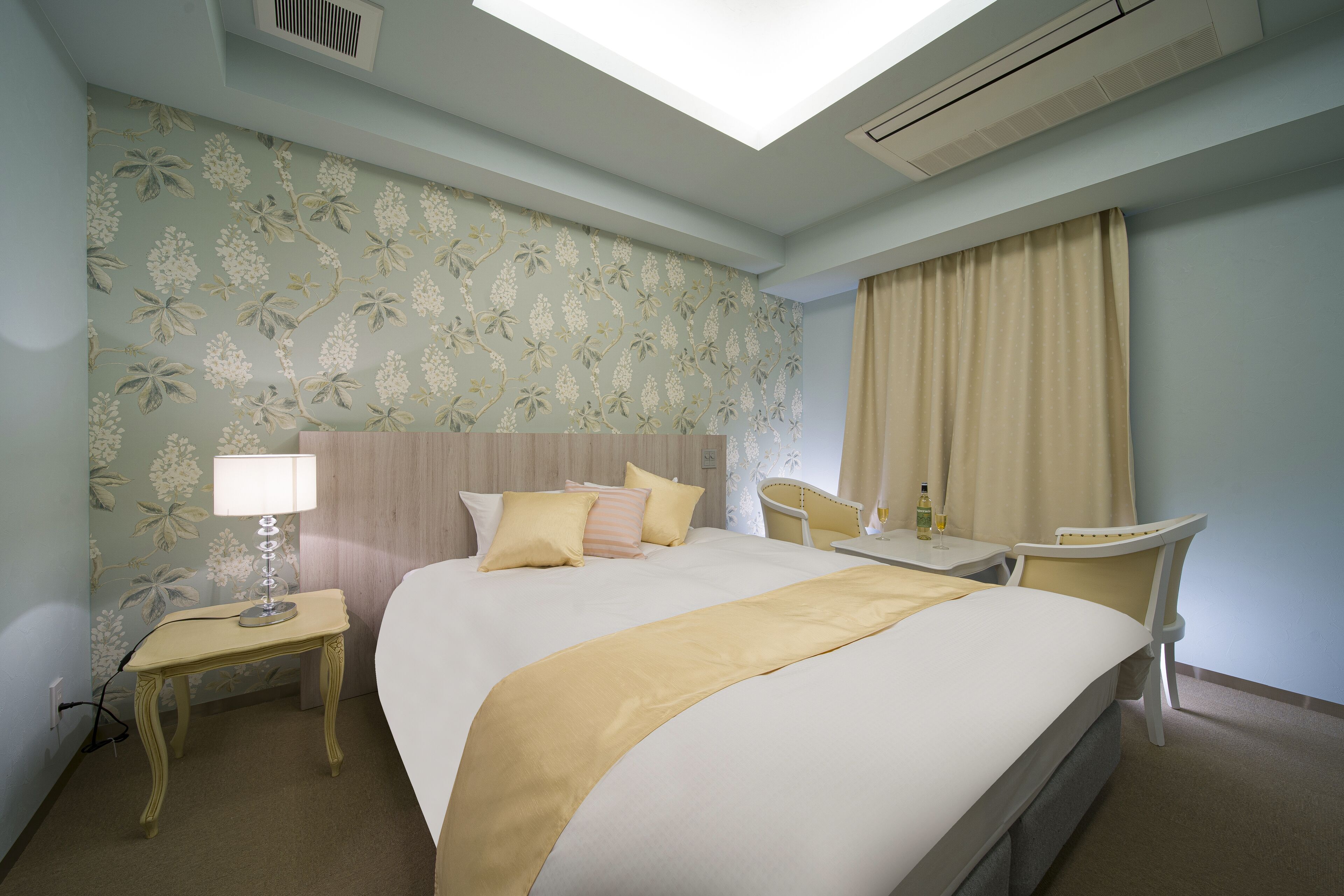 A hotel room with a bed, bedside table, lamp, chair, and a window with curtains.