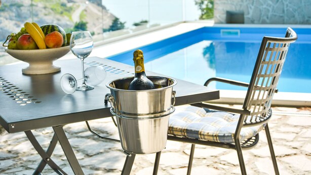 Una mesa con frutero, copa de vino y botella de champán, con vista a una piscina y al mar.