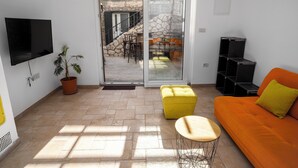 Una sala de estar con televisor, un pouf amarillo, un sofá naranja y una escalera que lleva a un espacio exterior.