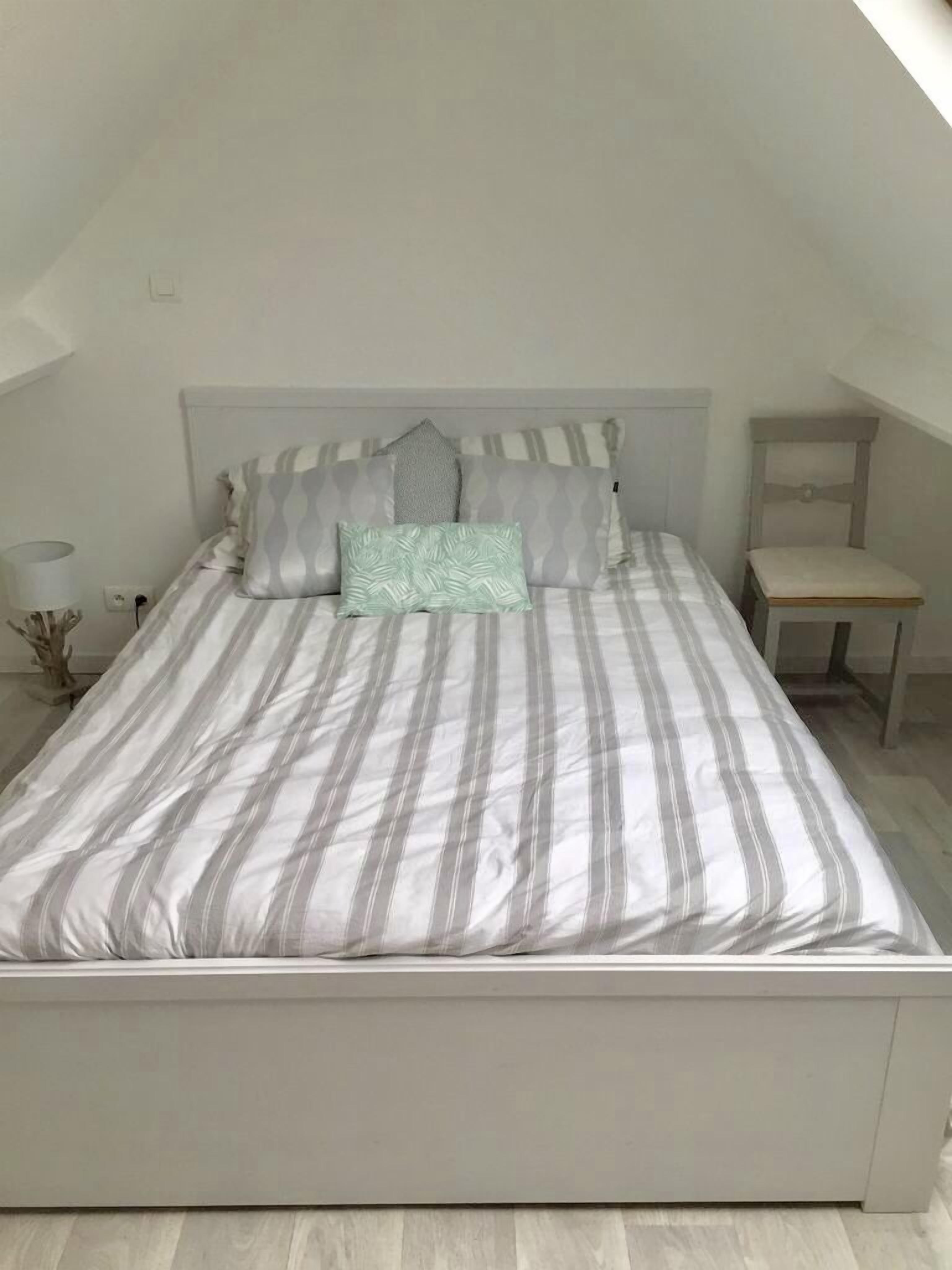 A neatly made bed with striped bedding and multiple pillows, a bedside table with a lamp, and a small wooden chair.