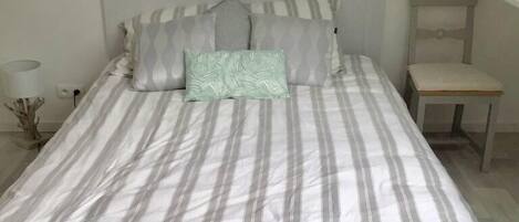 A neatly made bed with striped bedding and multiple pillows, a bedside table with a lamp, and a small wooden chair.