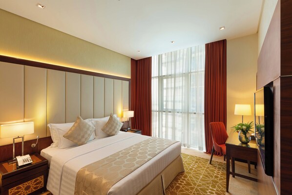 A hotel room with a large bed, bedside tables with lamps, a desk with a chair, and a large window with curtains. - Royal Continental Suites (Dubai)