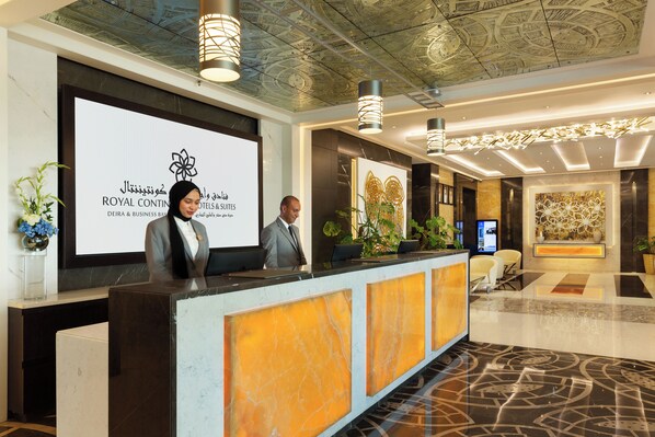 A hotel reception area with a modern design, featuring a reception desk, a large digital screen displaying the hotel name, and a decorative ceiling. - Royal Continental Suites (Dubai)