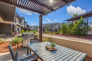A covered patio area with a wooden table, chairs, a grill, and a small garden.