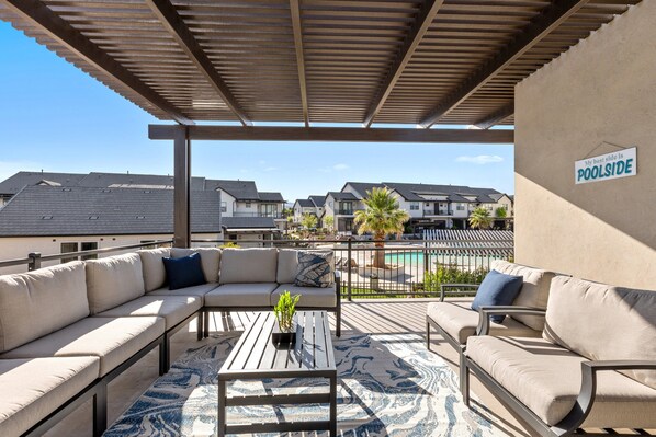 A covered outdoor seating area with a poolside sign, beige sofas, a wooden coffee table, and a patterned rug.
