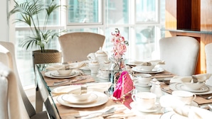 A dining table set with white plates, cups, and a pink vase with flowers, with a city view through large windows. - Elite Royal Apartment - Burj Khalifa & Fountain view - Senator (Dubai)
