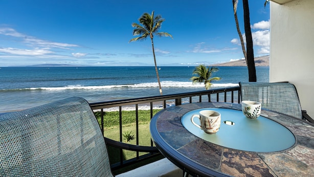 Un balcon avec une table et des chaises donnant sur une plage bordée de palmiers et où l’on peut admirer les vagues de l’océan.