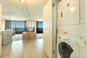 A laundry room with a front-loading washing machine, a small kitchenette, and a view of the balcony.
