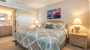 Une chambre à coucher avec un lit, des lampes de chevet, une table de nuit, un tableau au mur et une salle de bain visible à travers une porte ouverte.