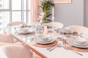 Ruokapöytä on katettu valkoisilla lautasilla, kulhoilla ja aterimilla, ja sen ympärillä on pehmustettuja tuoleja.