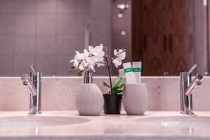 A bathroom sink area with two modern faucets, a potted plant, and a bottle of shampoo.