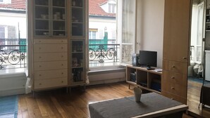 A room with a wooden floor, a table with a white object on it, a cabinet with glass doors, a television on a stand, and a window with a view of buildings and a balcony. - Monceau (Claude Pouillet) ID 302 (Paris)