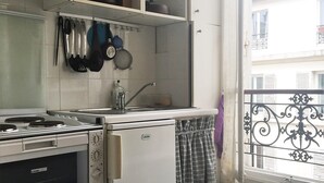 A compact kitchen with a stove, oven, and refrigerator. There are shelves above the counter holding various kitchen items. A window with a decorative iron railing is visible. - Monceau (Claude Pouillet) ID 302 (Paris)