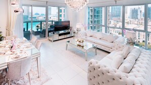 Ein geräumiges Wohnzimmer mit einem großen, weißen Sofa, einem Glastisch und einem Fernseher. Große Fenster bieten Blick auf die Stadt.