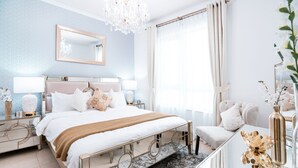 Ein Schlafzimmer mit einem Bett, Nachttischen, einem Spiegel, einer Chandelier und einem Fenster mit Vorhängen.