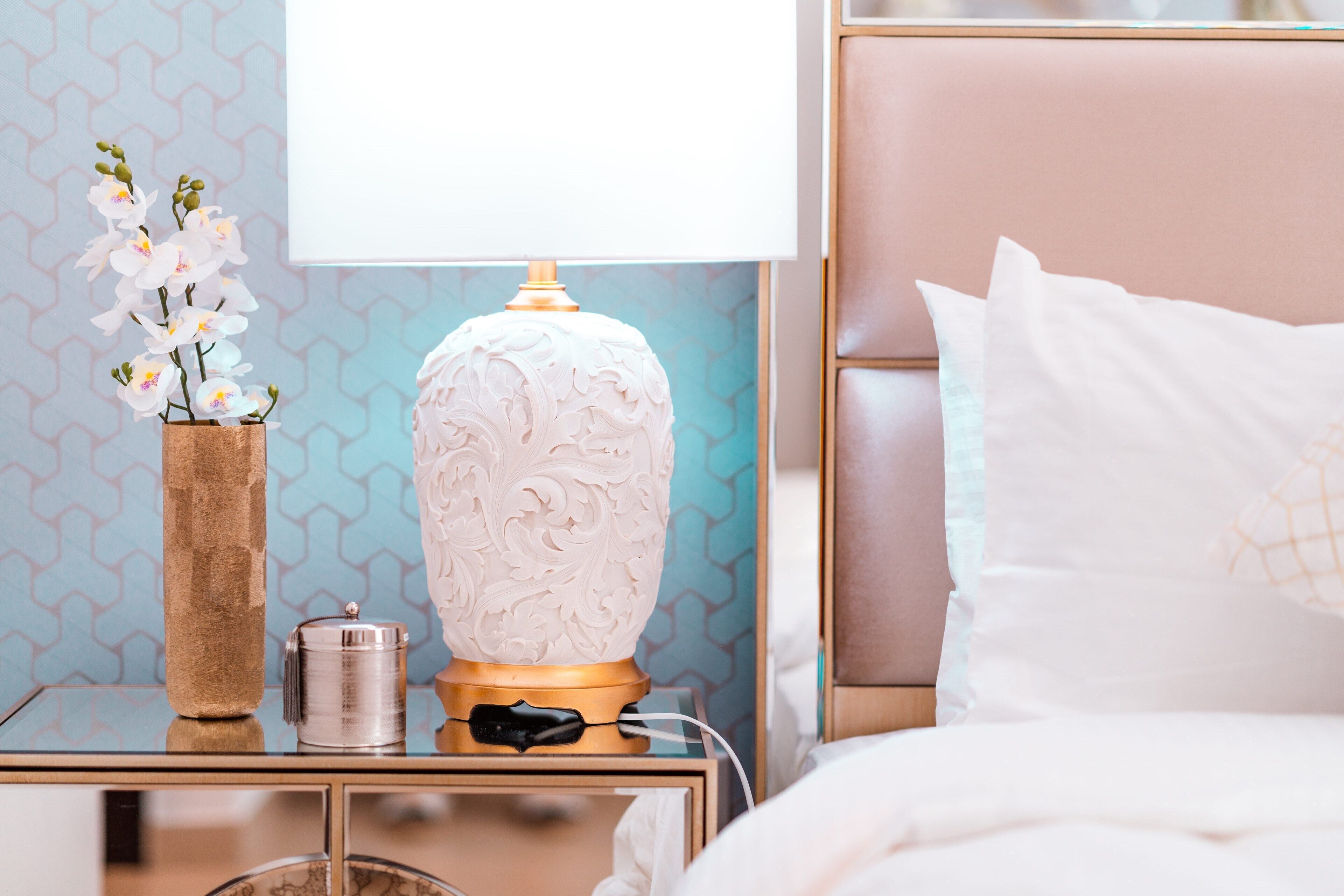 Een keurig opgemaakt slaapkamer met nachtkastje, een decoratieve vaas met bloemen, een lamp en een bed met kussens.