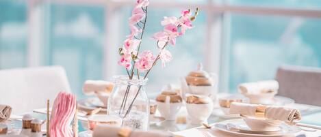 Een gedekte tafel met witte servies, roze bloemen in een glazen vaas en een goudkleurig lint als servet.
