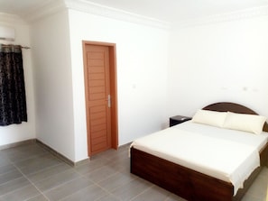A bedroom with a bed, a wooden door, a small bedside table, and a curtain. - Hôtel Naell Assinie (Assinie)
