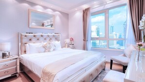 A modern bedroom with a large bed, a nightstand, a dresser, and a view of the city skyline through a large window.