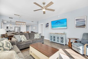 Ein geräumiges Wohnzimmer mit grauem Sofa, einem Holztisch und einem Deckenventilator. Im Hintergrund ist eine Küche mit weißen Schränken und einer Mikrowelle erkennbar.