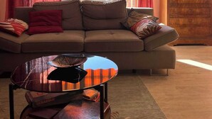 A living room with a sofa, a coffee table, and a view of buildings through a window with curtains. - COMFORTABLE 4/5-ROOM APARTMENT IN SECURE RESIDENCE WITH SWIMMING POOL (Saint-Raphaël)