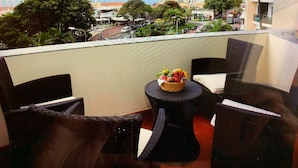 Un balcon agrémenté de meubles en osier, une petite table ronde avec un panier de fruits, et une vue sur un paysage urbain avec des bâtiments et des arbres.