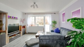 Ein modernes Wohnzimmer mit einem grauen Sofa, einem Flachbildfernseher, einem Holzschrank und einer großen, grünen Pflanze.