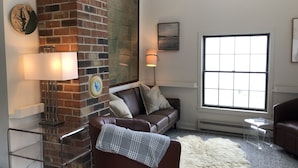 A living room with a brick fireplace, a brown leather sofa, a white shag rug, and a large window with a view.