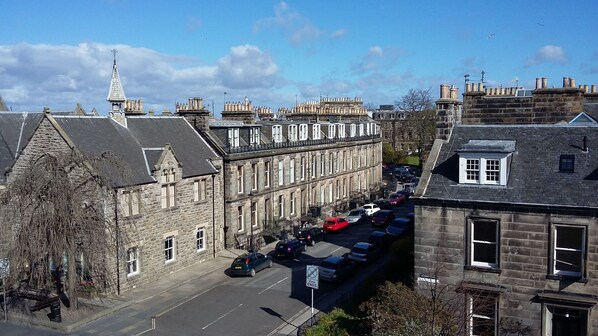 A street view with historic stone buildings, parked cars, and a clear sky.