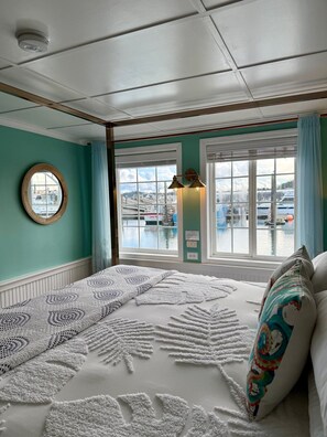 A room with a bed, a window with a view of boats, a round mirror, and a wall-mounted light fixture. - The Newport Belle (Newport)