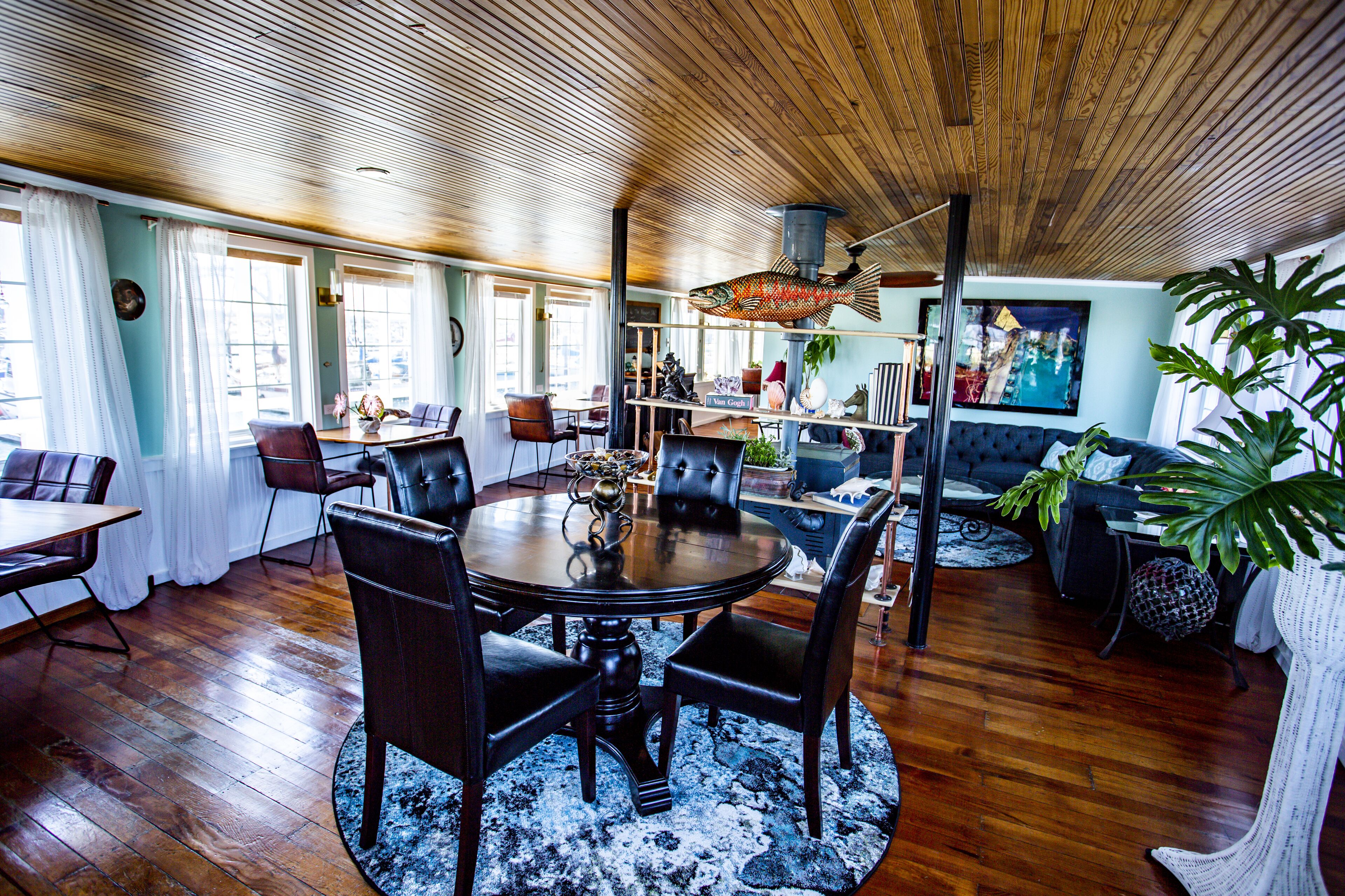 A dining area with a round table, chairs, and a blue rug. There is a bookshelf with various items and a large plant.