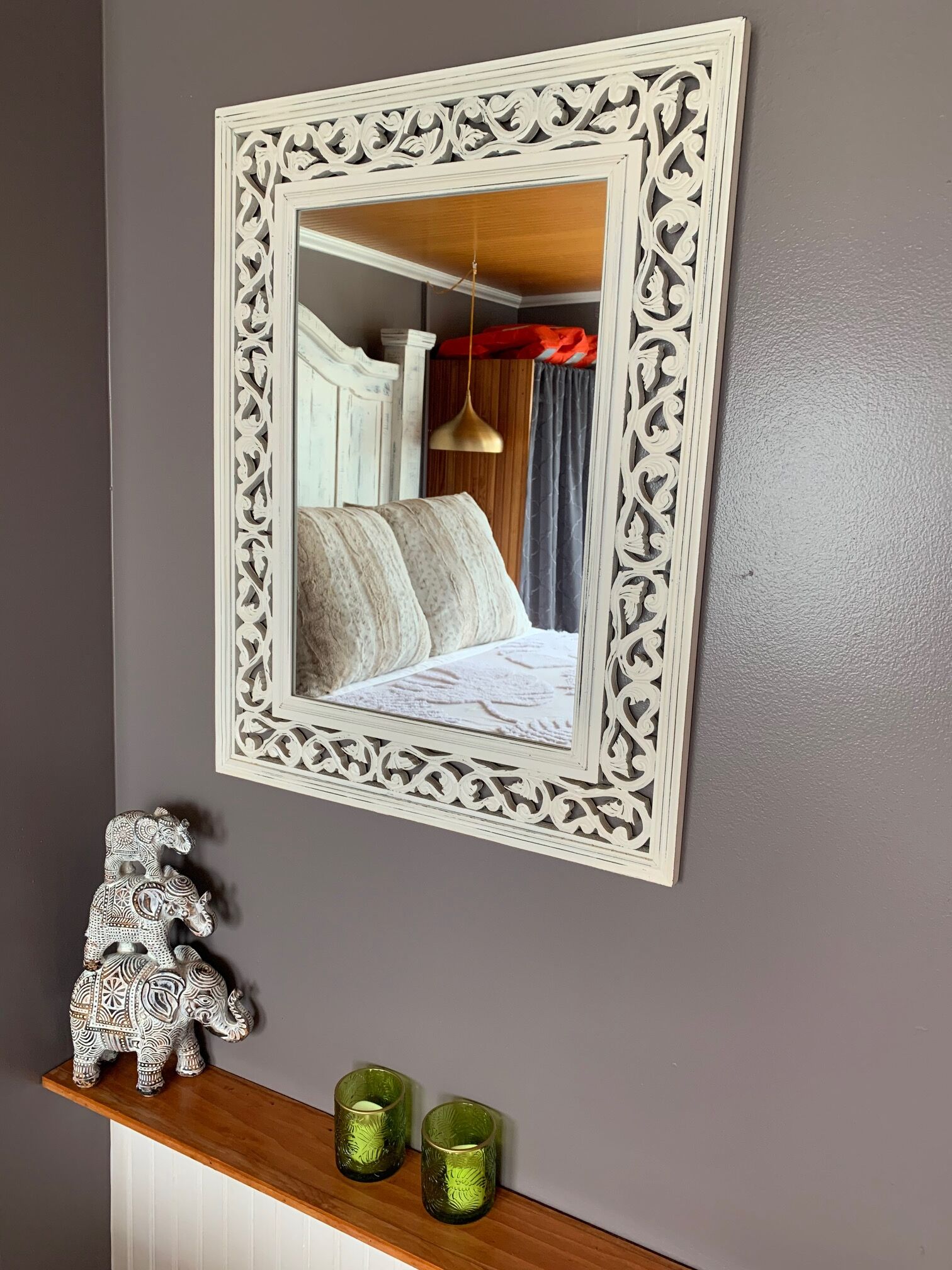 A mirror with an ornate white frame reflecting a bed with pillows and a hanging lamp. Below, a shelf holds decorative items and candles.