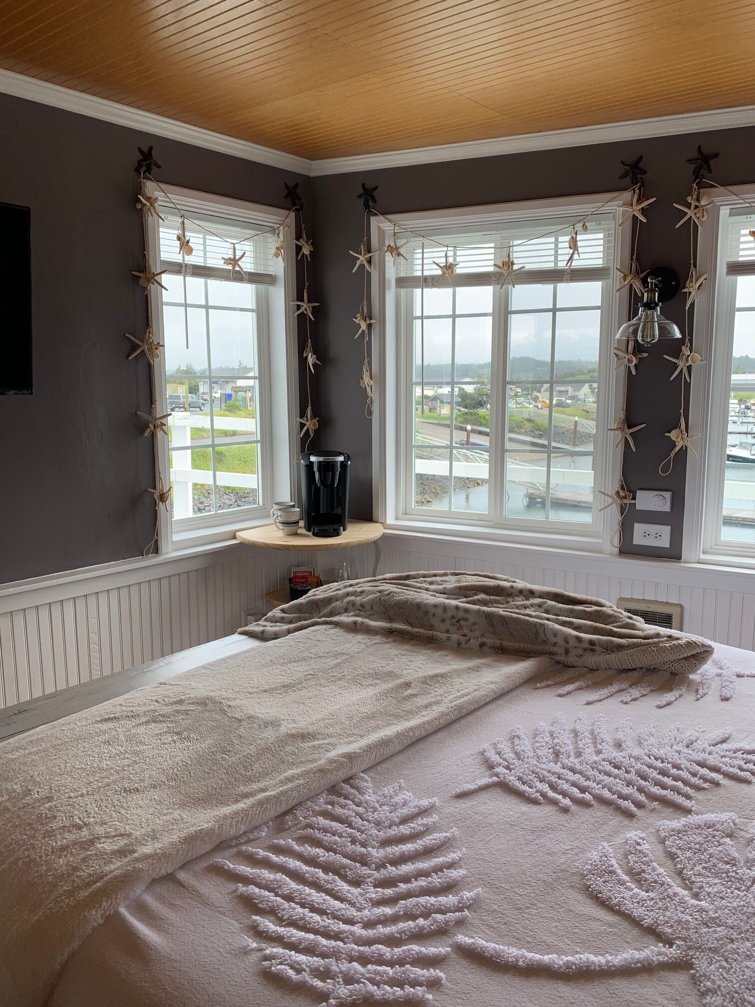 A bedroom with a large bed, two windows with starfish decorations, a coffee maker on a shelf, and a view of a marina.