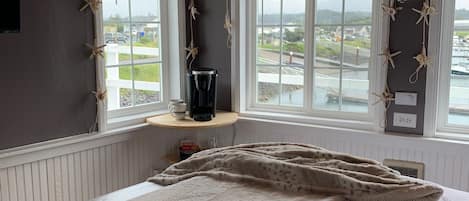 A bedroom with a large bed, two windows with starfish decorations, a coffee maker on a shelf, and a view of a marina.