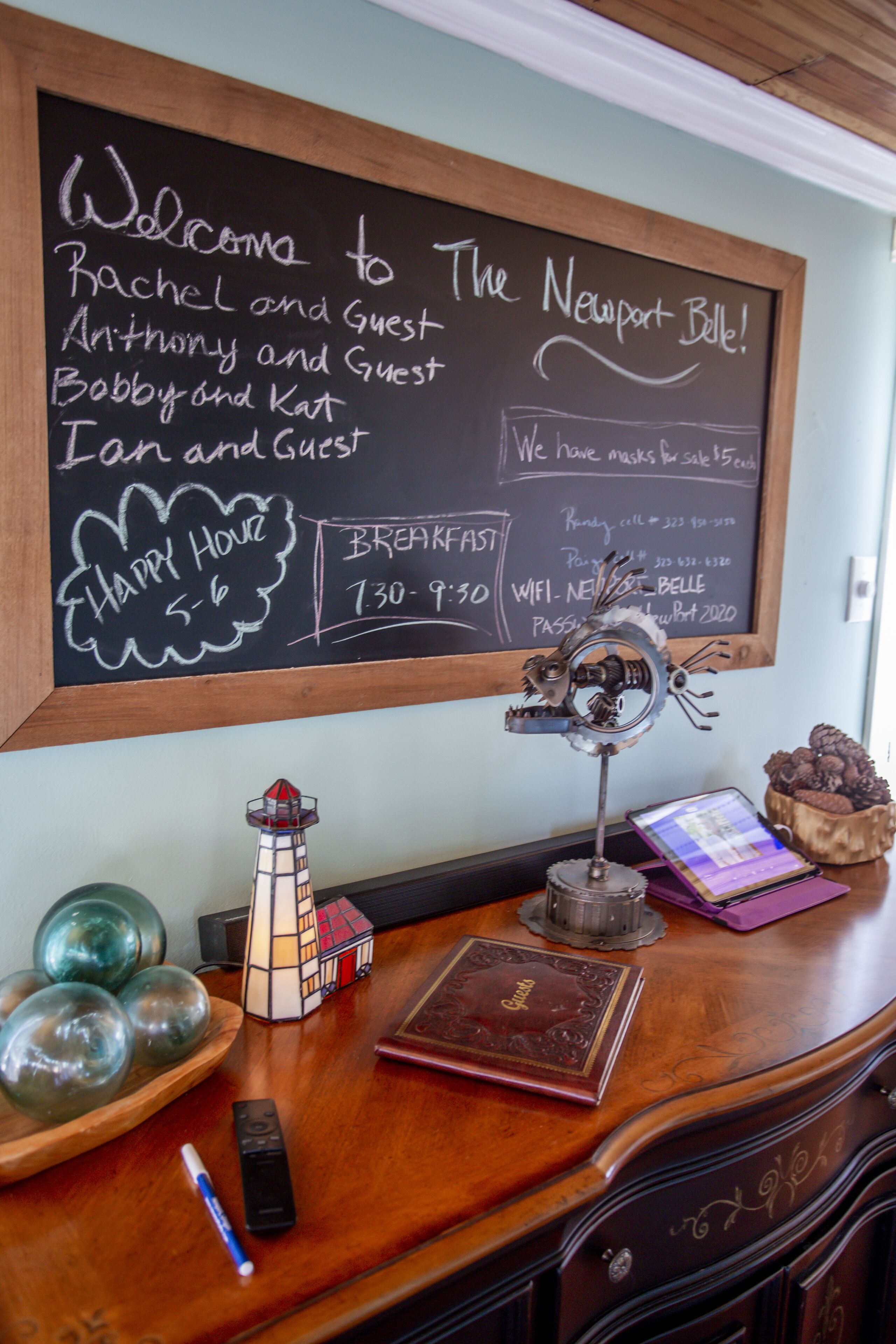 A chalkboard with a welcome message, a lighthouse model, a book, a remote, and decorative items on a wooden desk.