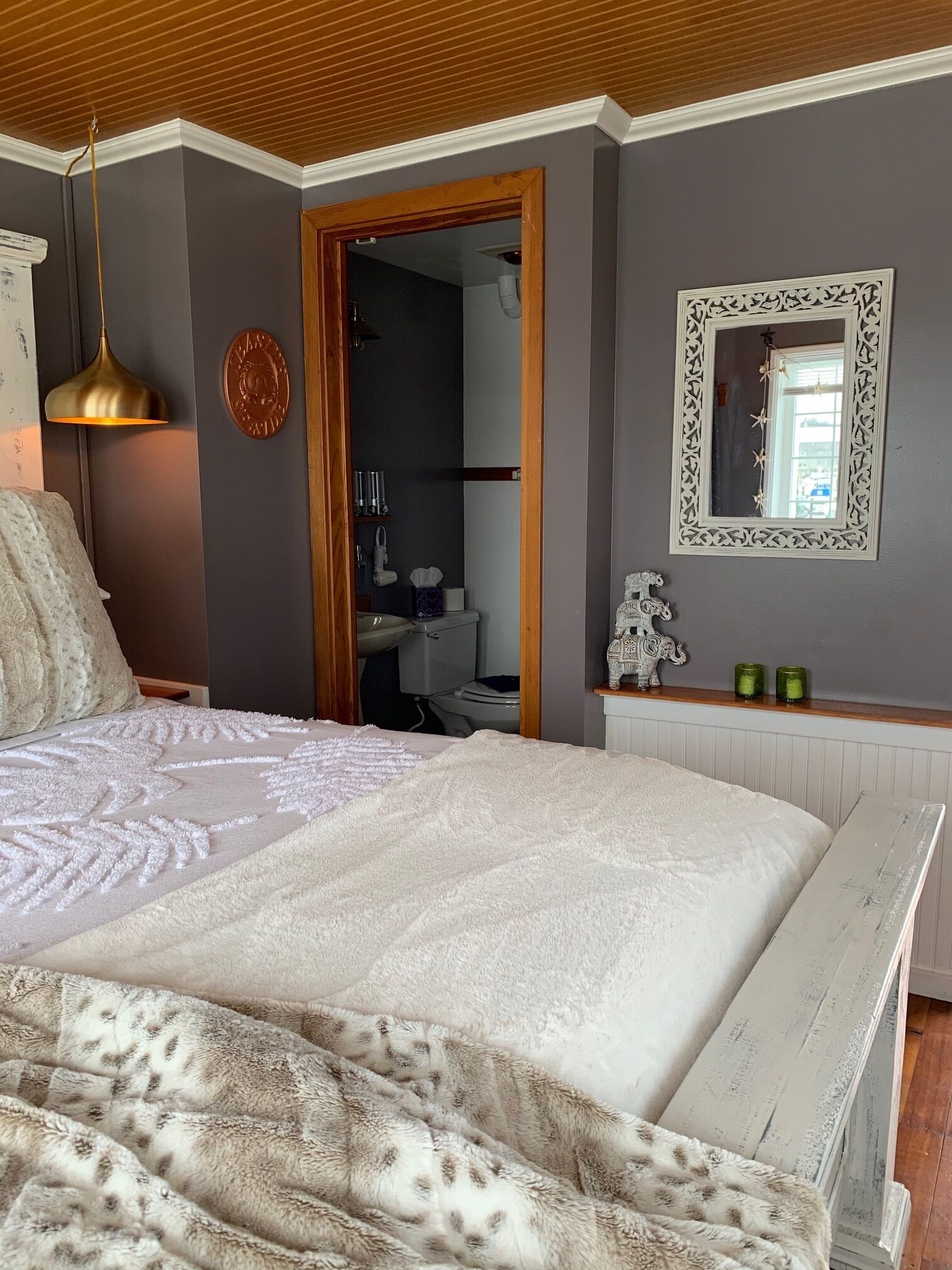 Une chambre à coucher avec un grand lit, un plafond en bois, un miroir et une salle de bain visible à travers une porte ouverte.
