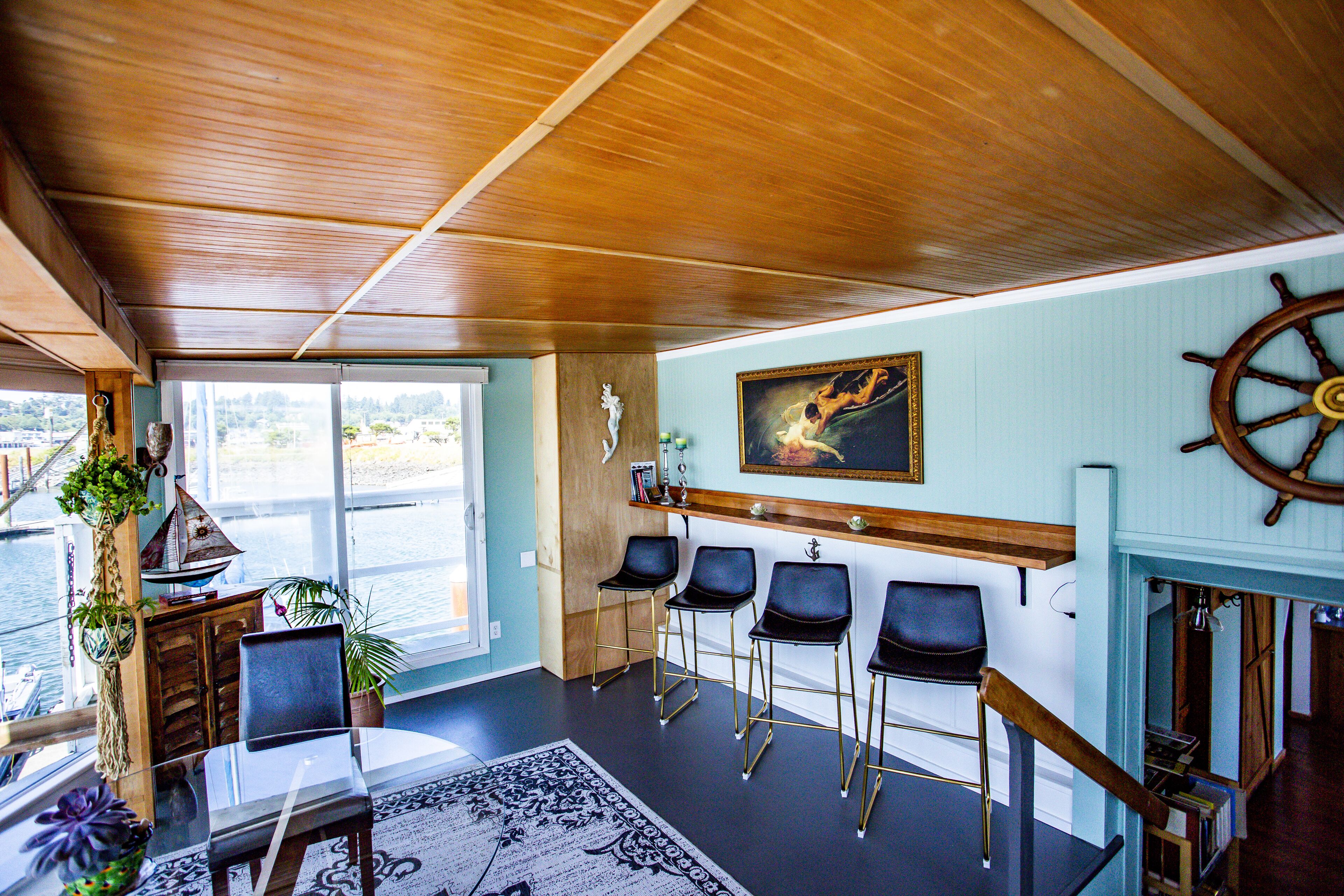 A room with a wooden ceiling, a large window with a view of the water, a sail wheel decoration, and a bar area with chairs.