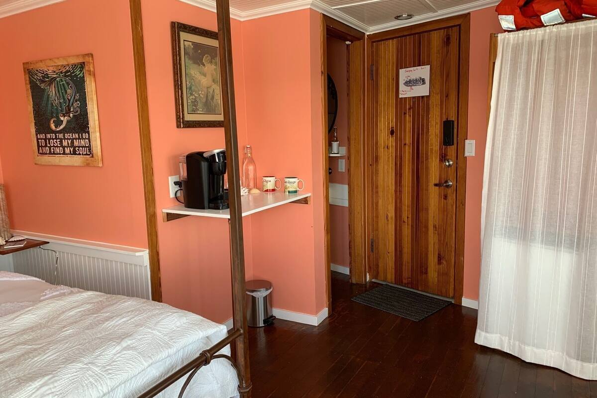 Ein Zimmer mit einem Bett, einer Kaffeemaschine, einem Regal mit Tassen, einer Tür und einem Vorhang.