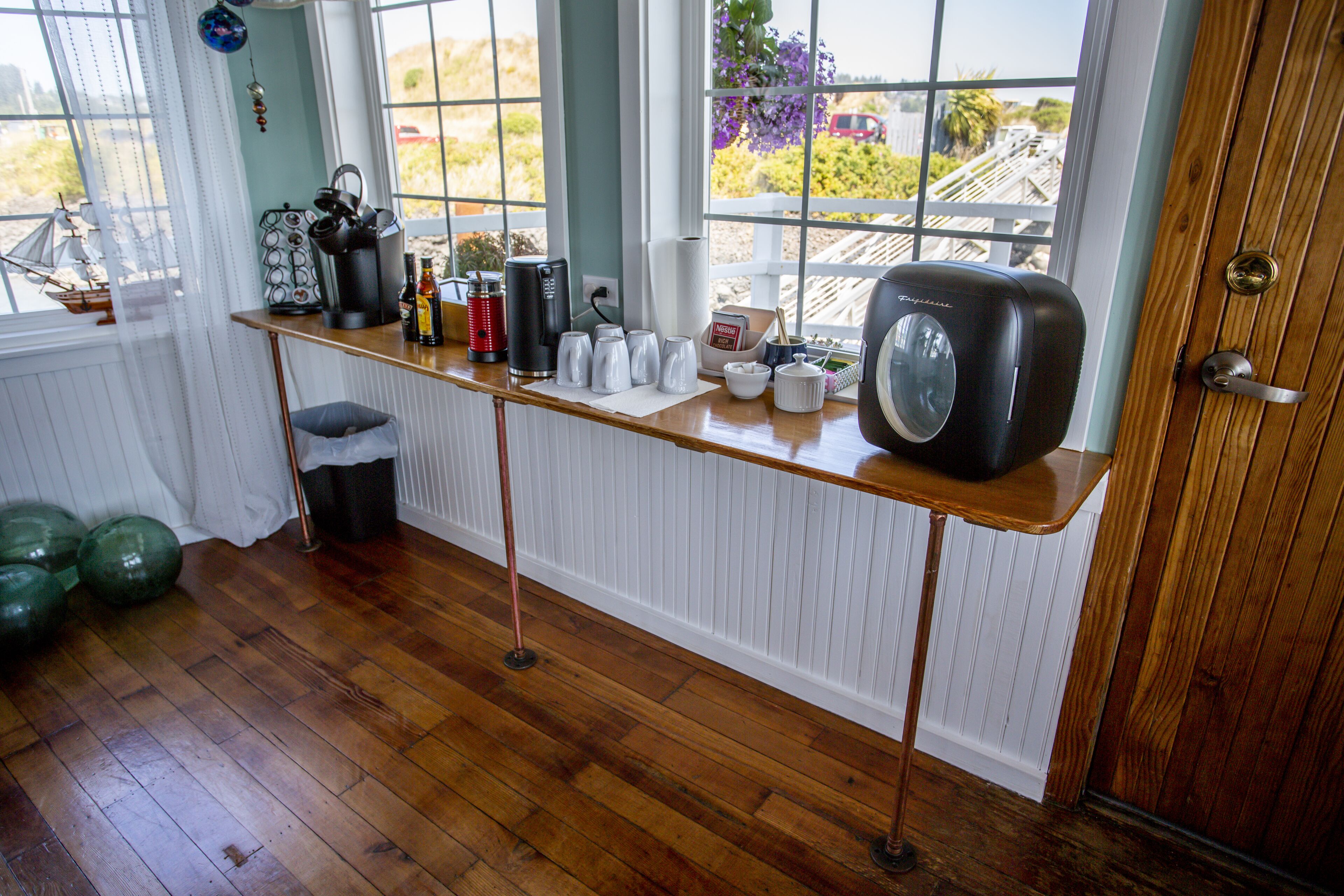 Una cocina con suelo de madera, encimera blanca y vistas al exterior a través de amplios ventanales.
