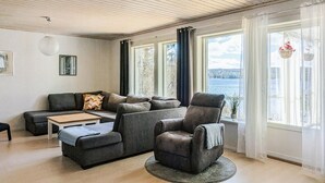 A living room with a sofa, armchair, and coffee table, large windows with curtains, and a view of a body of water. - Lovely home in Idkerberget (Idkerberget)