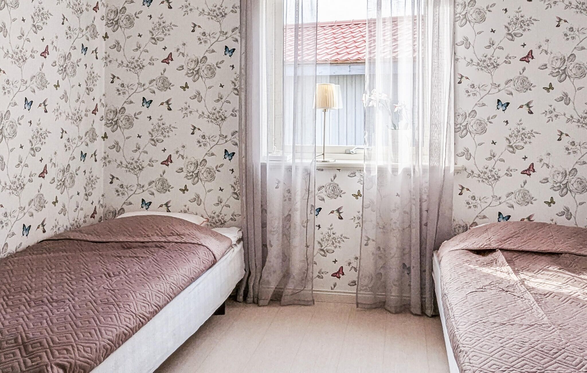 A room with two beds, floral wallpaper, and a window with sheer curtains.