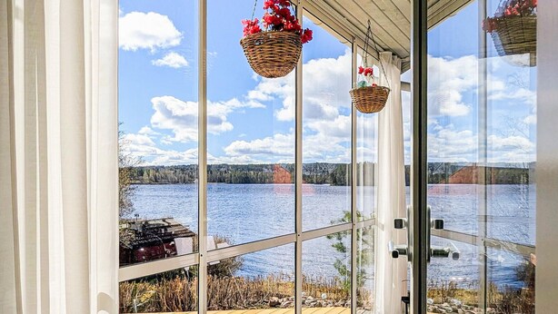 A room with large windows overlooking a lake, wooden deck, and hanging baskets with flowers. - Lovely home in Idkerberget (Idkerberget)