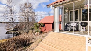 A wooden deck leading to a glass-enclosed building with a red roof, overlooking a body of water and trees. - Lovely home in Idkerberget (Idkerberget)