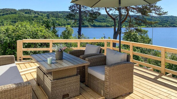 A wooden deck with wicker furniture, a table, and an umbrella, overlooking a lake and forested hills. - 3 bedroom lovely home in Strömstad (Strömstad)
