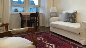 Una sala de estar con un sofá blanco, un tapete estampado, una mesa de comedor de madera con sillas y un ventanal con vista a edificios.