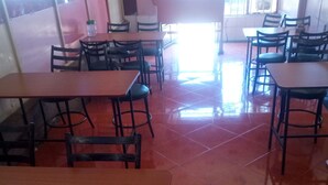 Un espace repas avec plusieurs tables et chaises, un sol carrelé et une grande fenêtre lumineuse à l’arrière-plan.